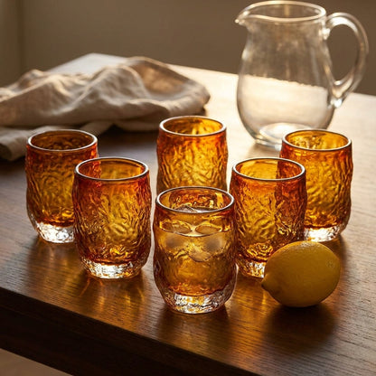 Seis copos Marbella Glow em vidro ambar numa mesa de madeira escura ao fim da tarde, um com agua e gelo