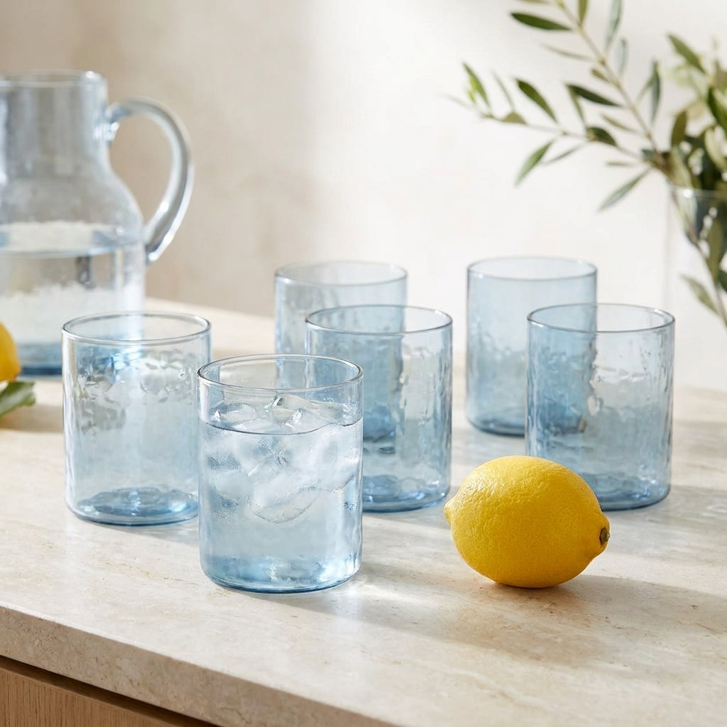 Seis copos altos martelados Azure em vidro azul-claro numa bancada de pedra, um com agua e gelo