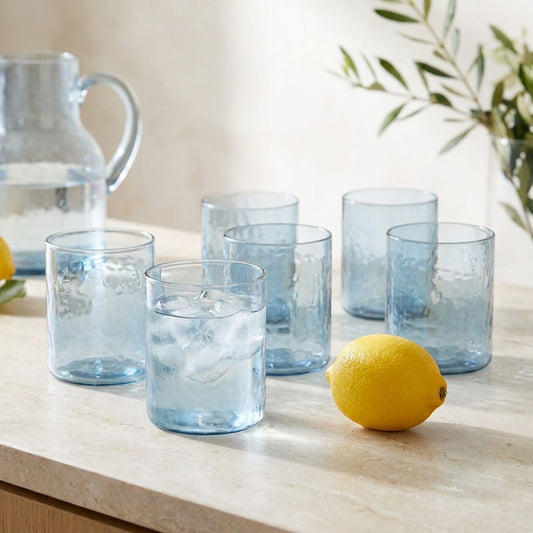 Seis copos altos martelados Azure em vidro azul-claro numa bancada de pedra, um com agua e gelo
