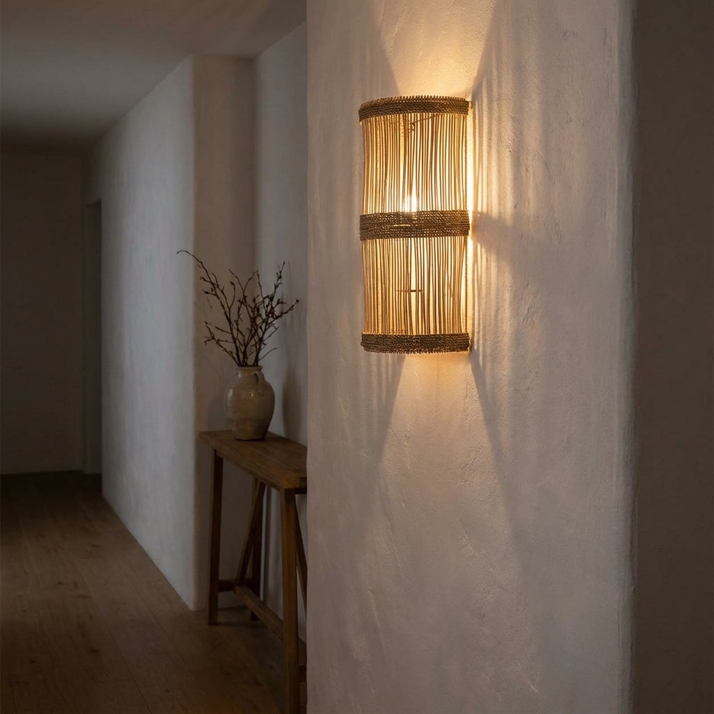 Aplique de parede Jalal em rattan natural aceso num corredor, com riscas de luz e sombra na parede caiada
