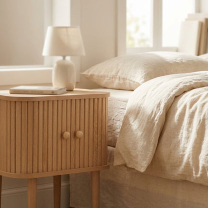 Mesa de apoio Nikko em madeira natural com portas em ripas curvas, ao lado de uma cama de linho cru
